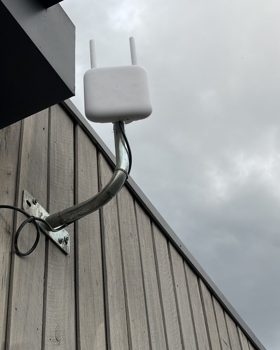 A white outdoor WiFi unit with two short antennas on a galvanised arm bolted to timber wall cladding