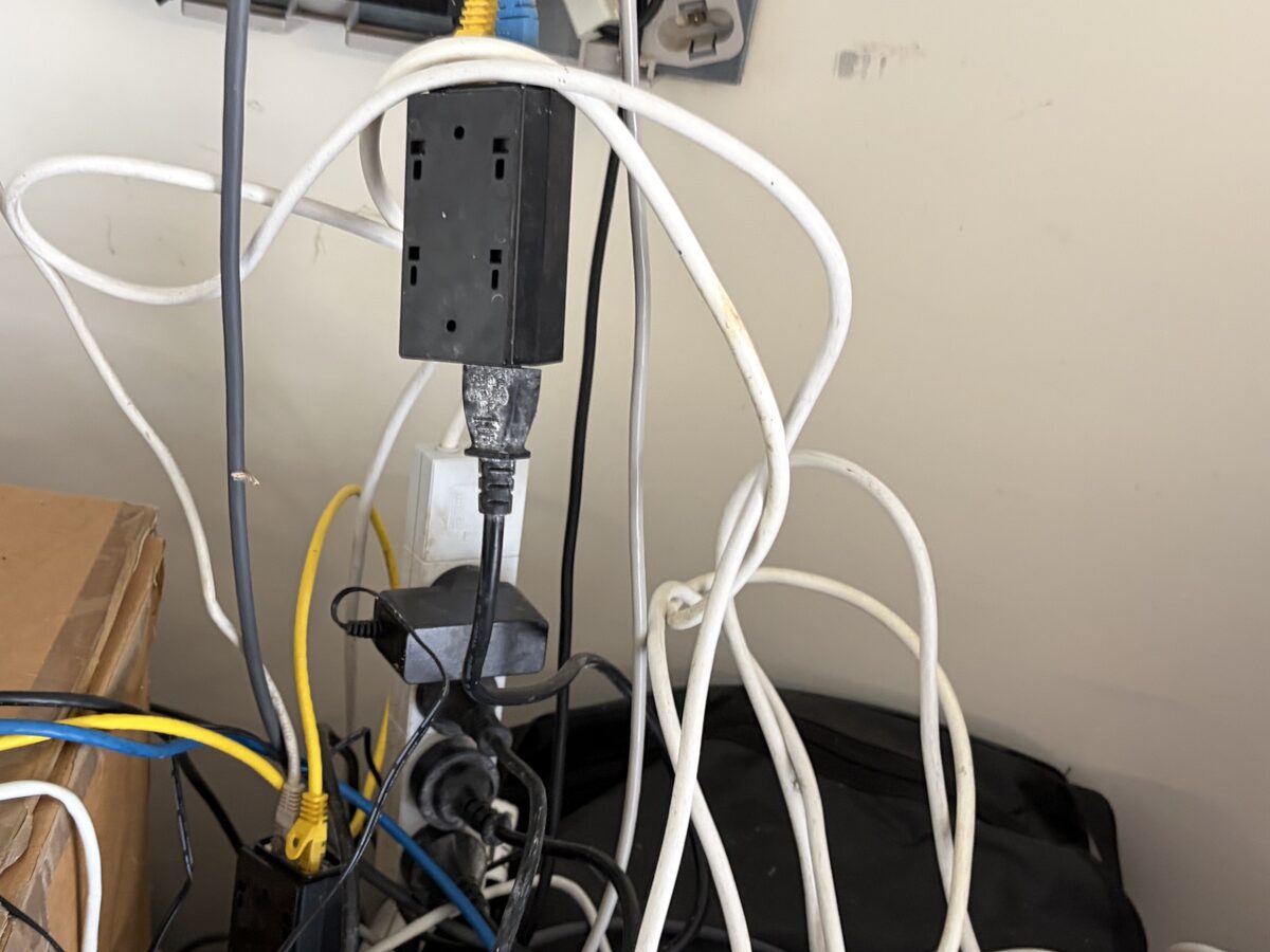 A power board hanging in mid-air by its own lead, with plug packs, a second power board and a tangle of white, yellow and blue cable below it
