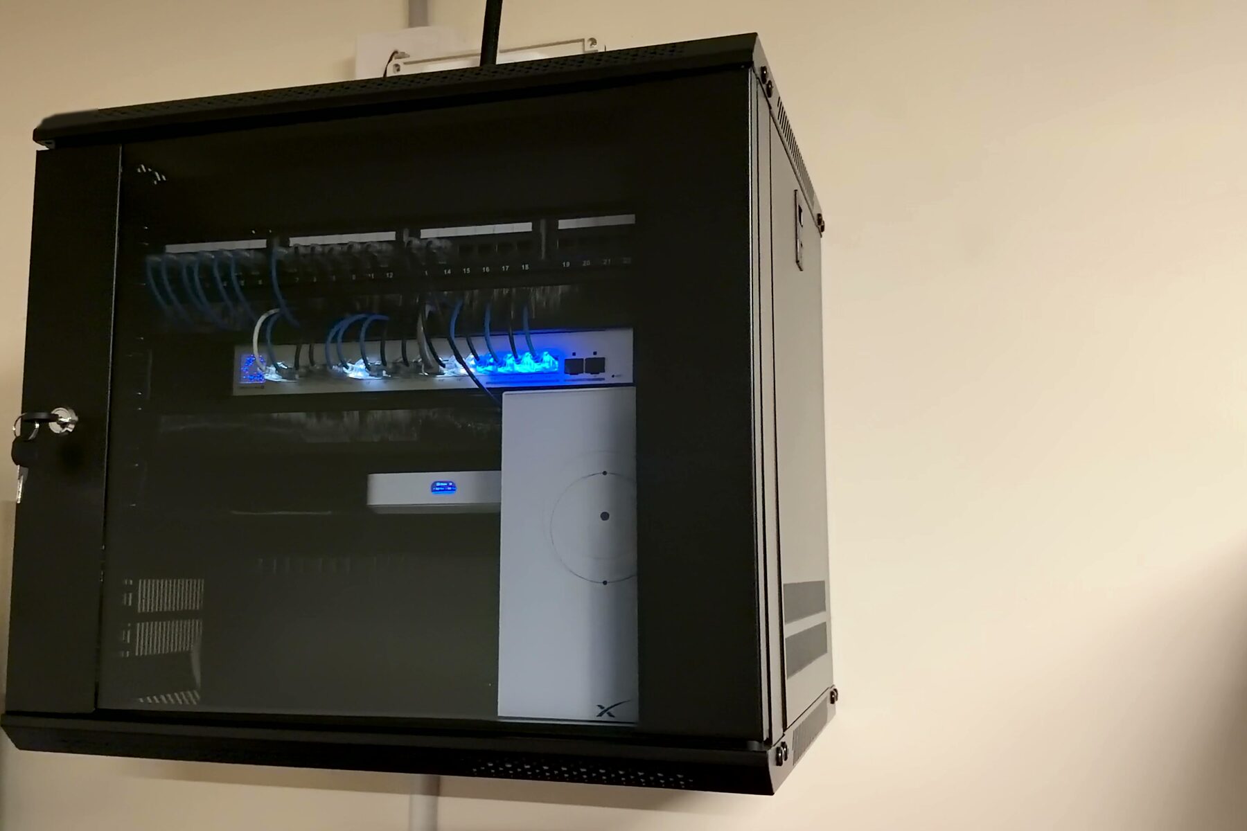 A wall-mounted network cabinet with a glass door, holding a patch panel, a lit network switch and short blue patch leads